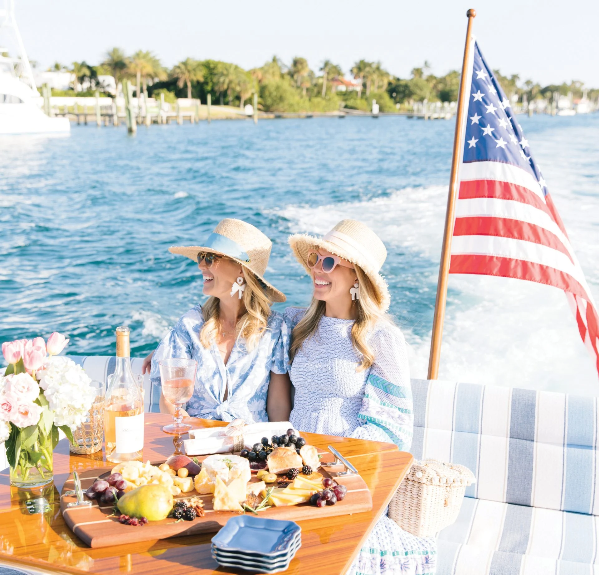 Two women enjoying a boat ride on open water, wearing sun hats and smiling as an American flag waves behind them.